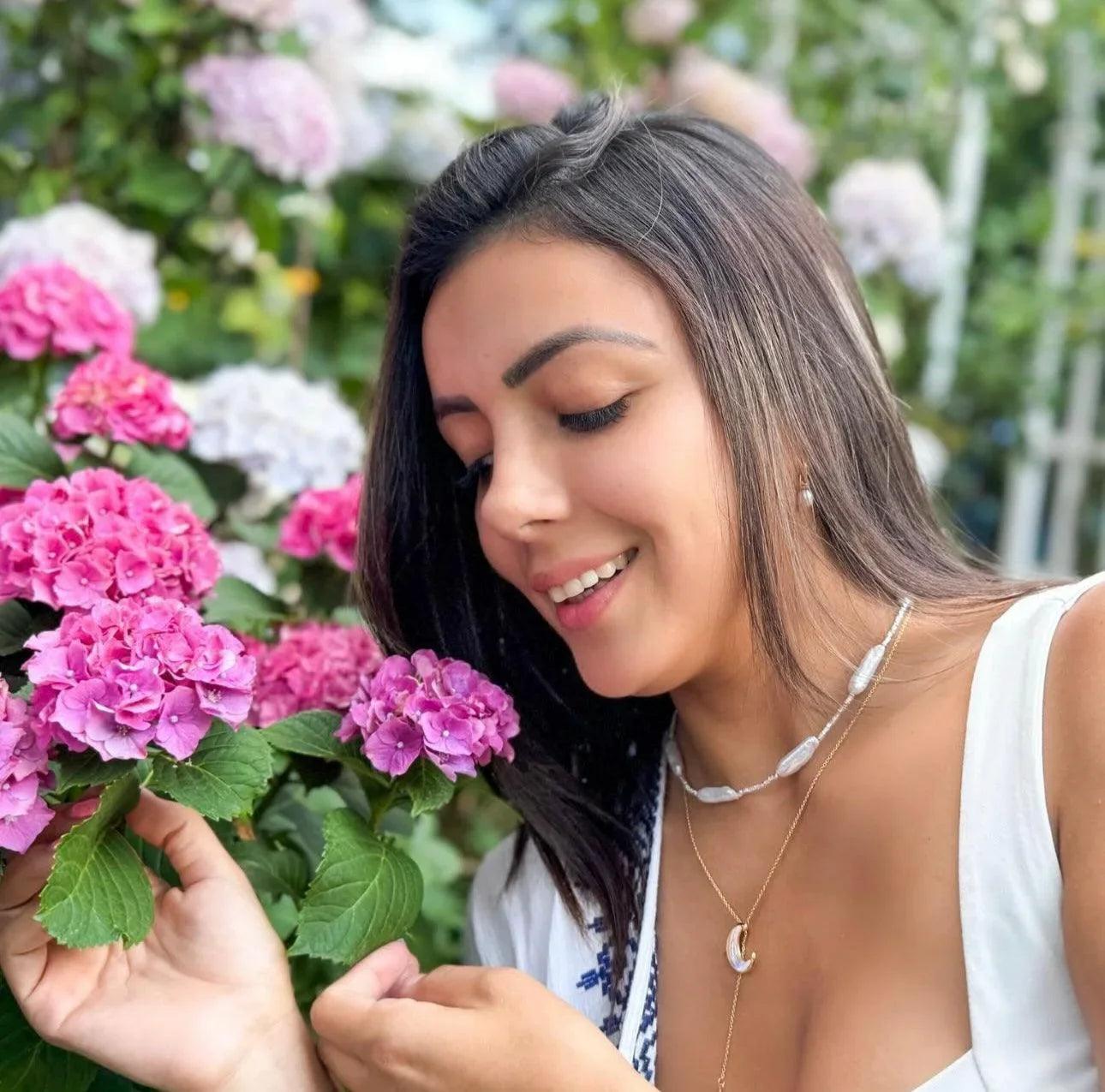 Smiling woman with dark hair holding pink hydrangea flowers in a lush garden setting