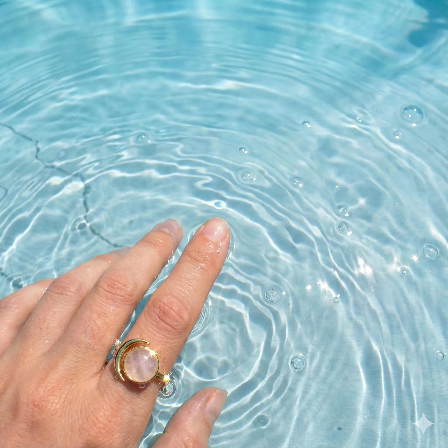 Hand with gold ring featuring round gemstone touching clear blue water with ripples
