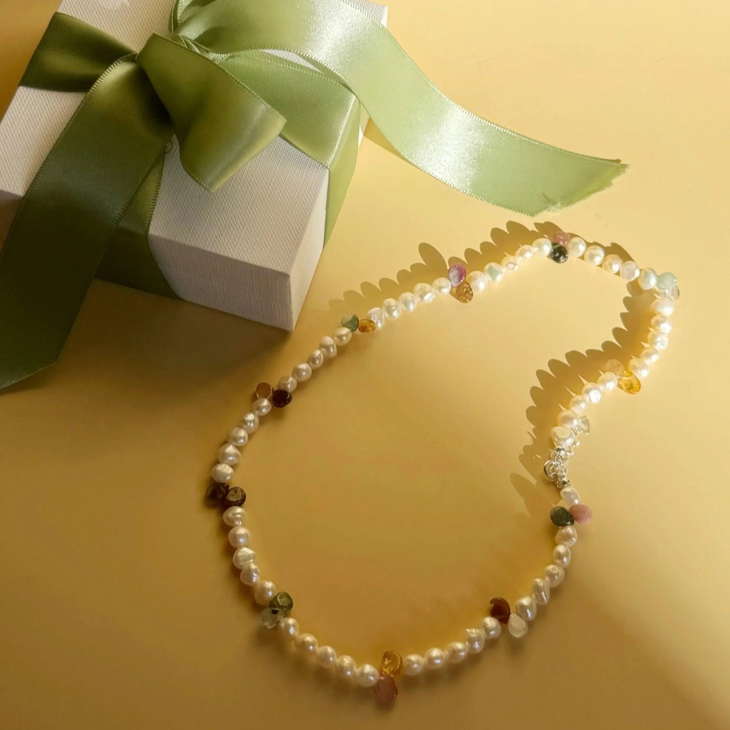 Pearl necklace with multicolor gemstone accents next to gift box with green ribbon on beige surface