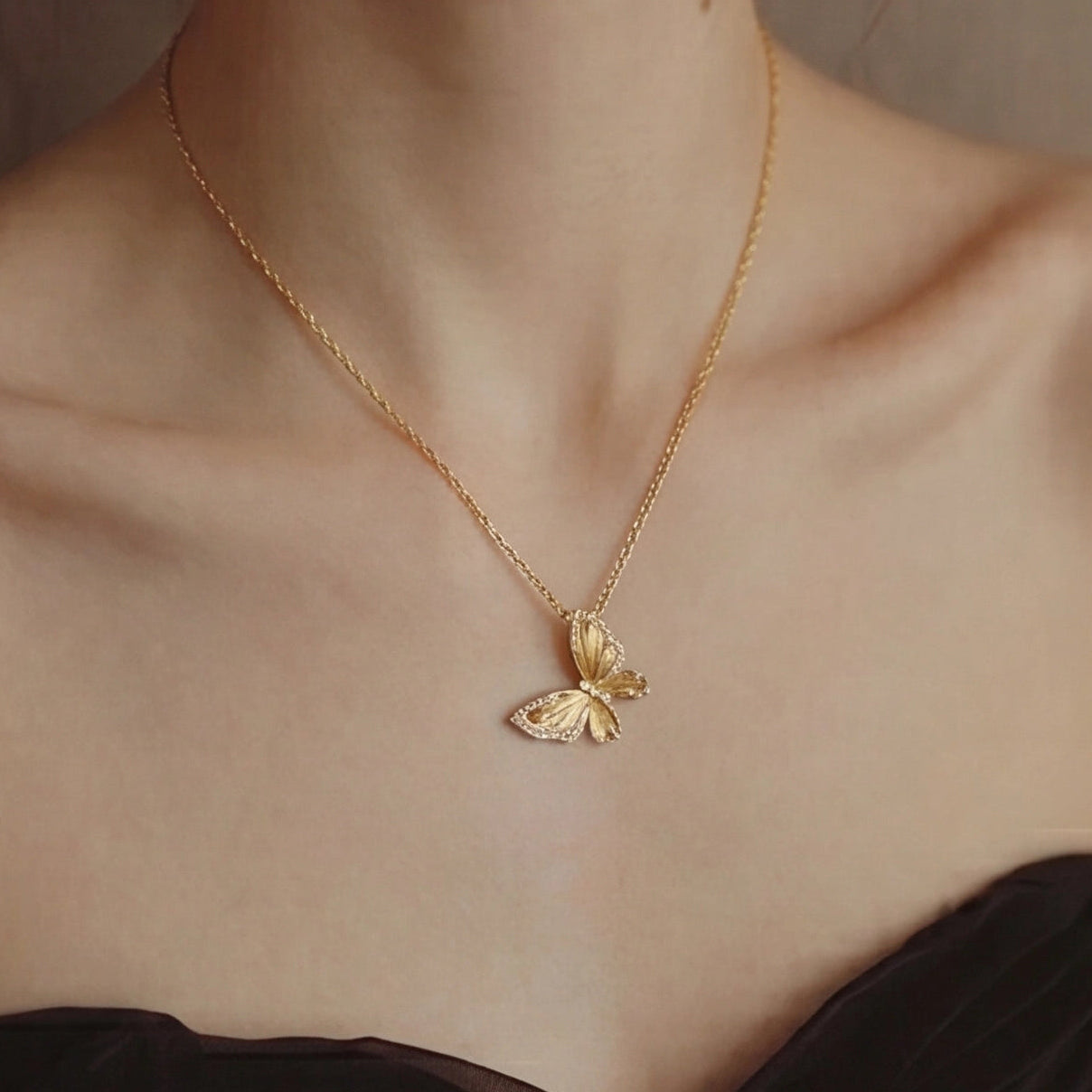 Gold butterfly necklace on a person wearing a black top against a neutral background