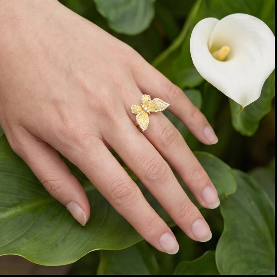 Hand wearing a gold ring with a butterfly design, surrounded by green leaves and a white flower.