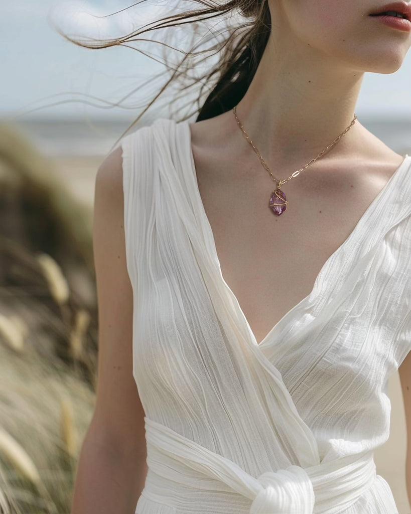 Woman wearing white pleated dress and gold chain necklace with purple gemstone pendant at beach
