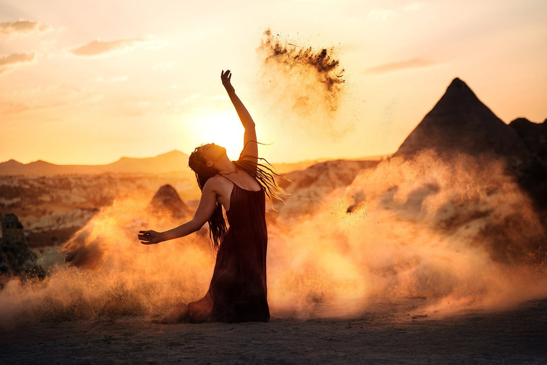 Woman in flowing dress dancing with sand at sunset in desert landscape with rock formations