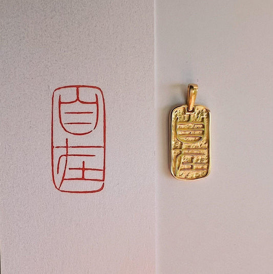 Gold pendant necklace with embossed calligraphy design next to red calligraphy symbol on paper