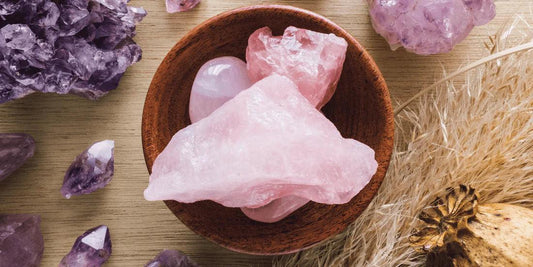 Various rough rose quartz and amethyst crystals on a wooden surface with dried grass and wooden bowl