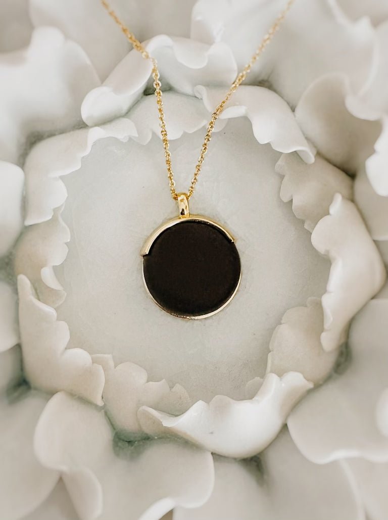 Gold necklace with round black pendant placed on white flower petals, elegant jewelry close-up