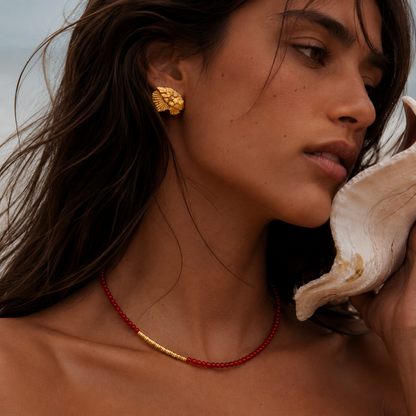 Woman wearing gold earrings and a red beaded necklace, holding a shell to her ear.