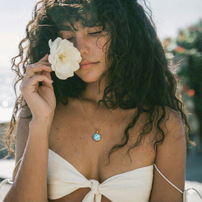 Woman in white top holds white flower near face, wearing larimar pendant necklace with gold chain