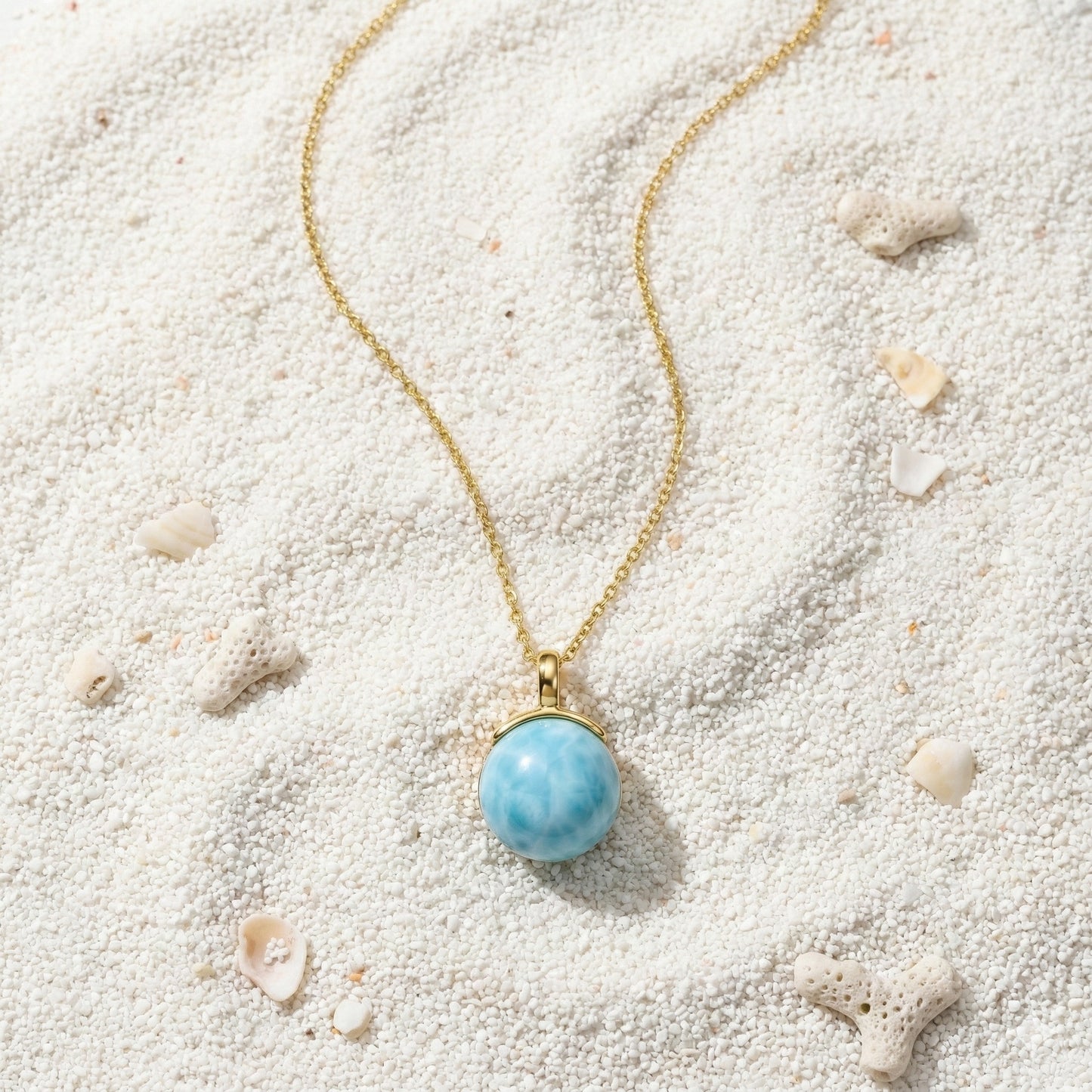 Gold chain necklace with a round larimar pendant on white coral sand with small seashells