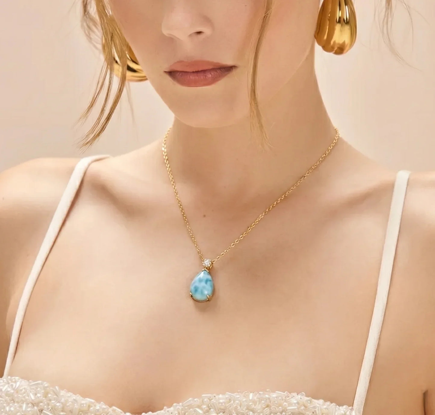 Woman wearing a gold necklace with a blue pendant against a neutral background