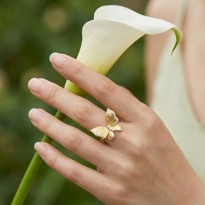 Hand wearing a gold butterfly ring holding a white flower with a blurred green background