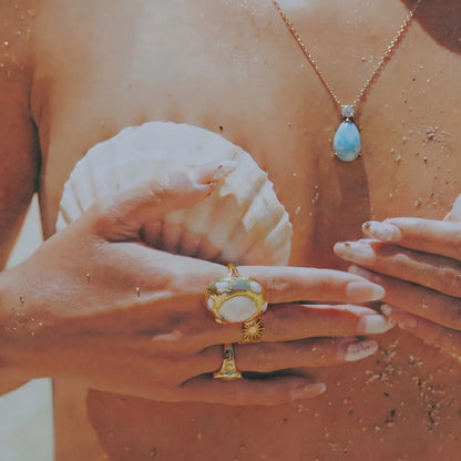 Close-up of hands wearing gold rings with a shell and necklace in the background